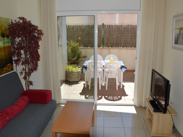 Una sala de estar con un sofá, una mesa de centro, un televisor sobre un mueble bajo y un ventanal corredizo que da a un patio exterior con una mesa y sillas.