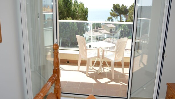 Een balkon met een houten stoel, een tafeltje en uitzicht op zee en gebouwen.