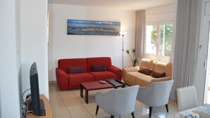 Een woonkamer met een rode bank, een beige bank, een salontafel en twee fauteuils. Er is een flatscreen televisie op een standaard, een schilderij aan de muur en een vloerlamp.