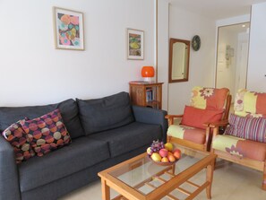 Un salon avec un canapé, un fauteuil et une table basse en verre sur laquelle sont disposés des fruits.