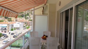 Un balcón con toldo a rayas, sillas blancas, una mesa con fruta y vistas a una calle con coches y edificios.