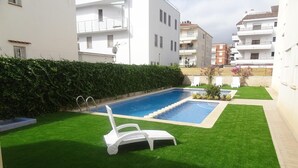Una piscina con un pequeño solárium, rodeada de césped cuidado y un seto.