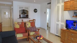 Un salón con un sofá, sillas, una mesa de centro con fruta, un televisor y una cocina al fondo.