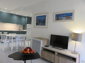 A modern kitchen with white cabinets, a dining area with white chairs, a TV on a wooden stand, and artwork on the wall.
