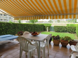 Une terrasse extérieure dotée d’un auvent à rayures, d’une table sur laquelle se trouve un panier de fruits, de chaises en plastique blanches et de plantes en pot.