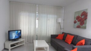 A living room with a flat-screen TV, a white coffee table, a grey sofa with orange cushions, and a painting of red flowers on the wall.