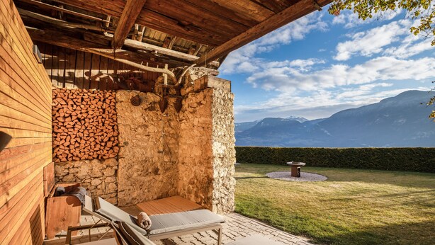 Un muro de piedra con un banco de madera y sillones de descanso, una pila de leña y vistas a montañas.