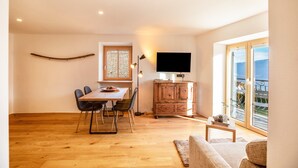 Un área de comedor con una mesa y sillas de madera, un televisor sobre un mueble bajo y vistas al exterior a través de ventanas amplias.