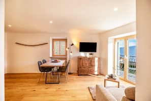 Un área de comedor con una mesa y sillas de madera, un televisor sobre un mueble bajo y vistas al exterior a través de ventanas amplias.