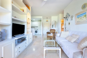 A living room with a flat-screen TV, white furniture, a glass coffee table, and a sofa with patterned cushions.