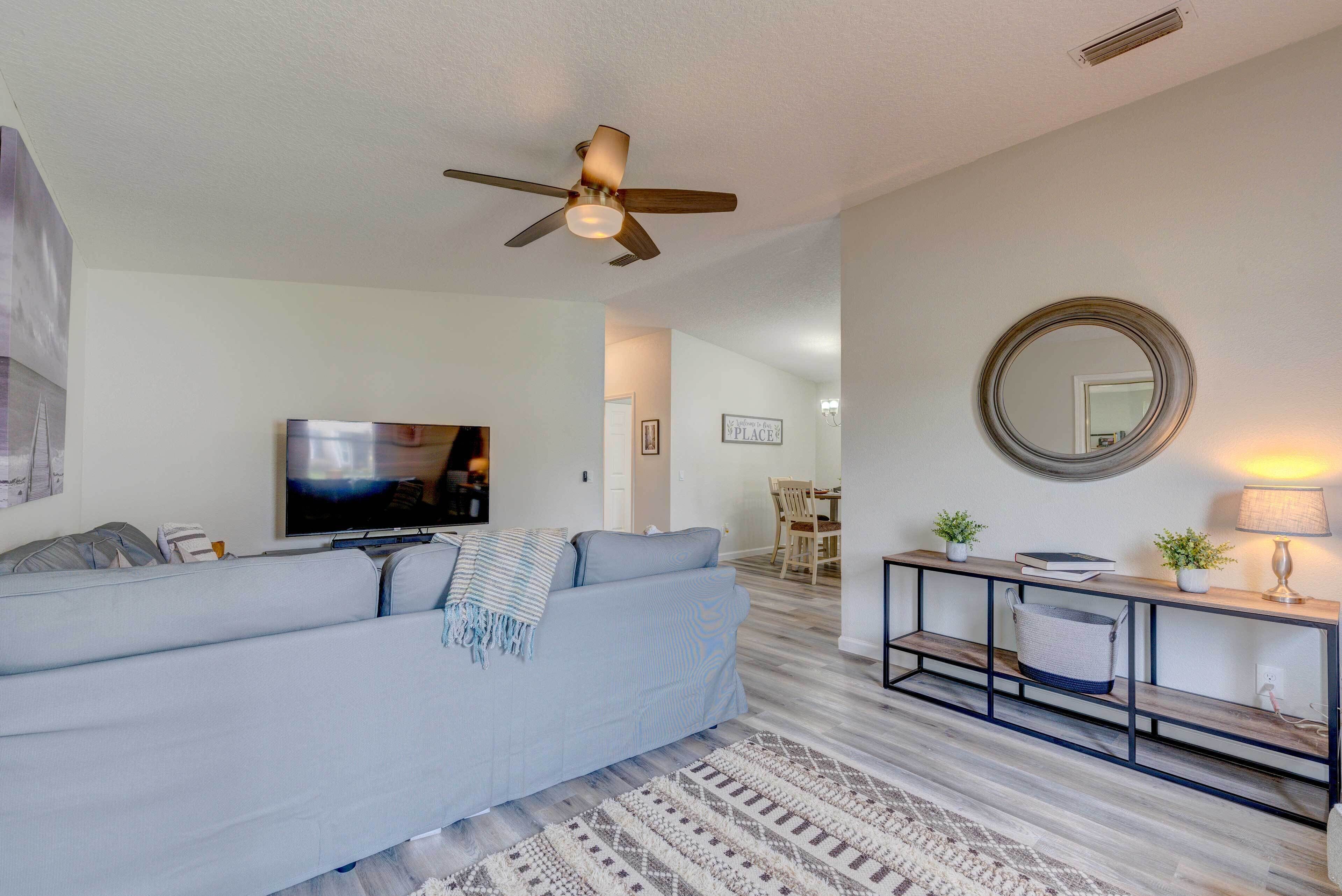 A modern living room with a light blue sofa, a flat-screen TV, a ceiling fan, a wooden console table with a lamp, and a mirror on the wall.