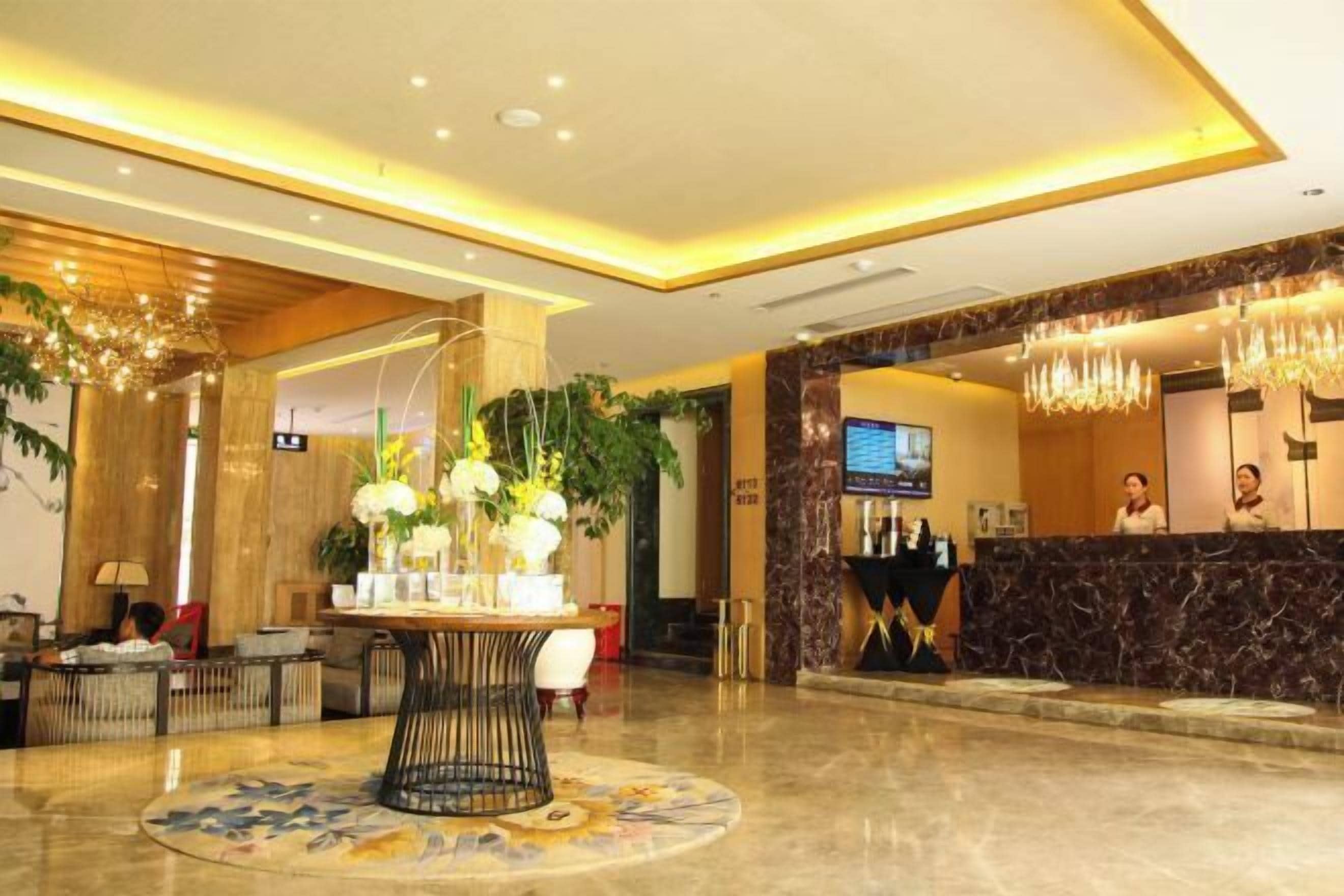A hotel lobby with a reception desk, a round table with a vase of flowers, and a chandelier.