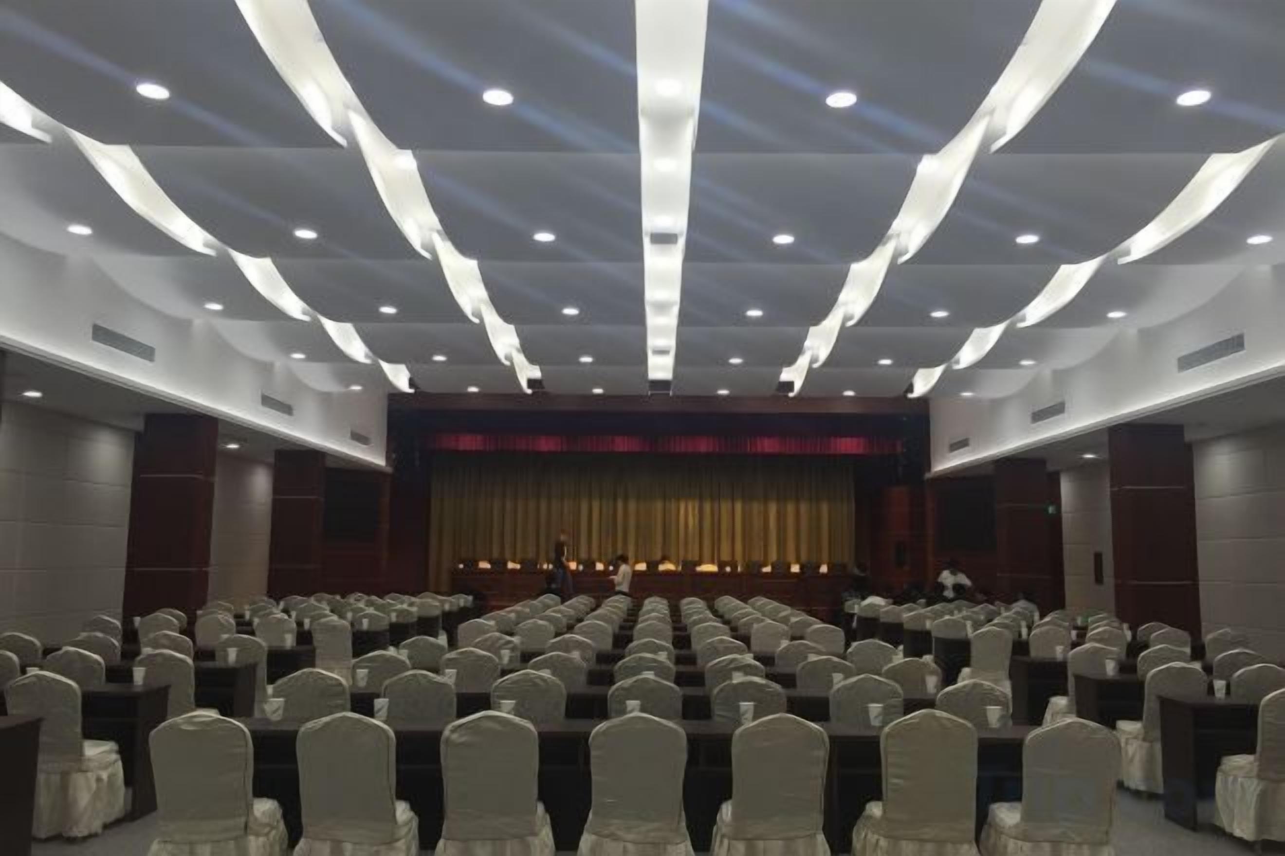 Un salón de conferencias con filas de sillas, un escenario con cortina y una persona de pie al frente.