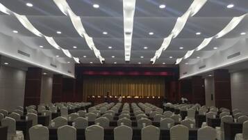 Un salón de conferencias con filas de sillas, un escenario con cortina y una persona de pie al frente.