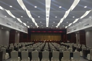 A conference room with rows of chairs, a stage with a curtain, and a person standing at the front.