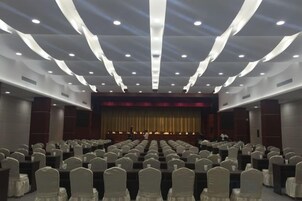 A conference room with rows of chairs, a stage with a curtain, and a person standing at the front.