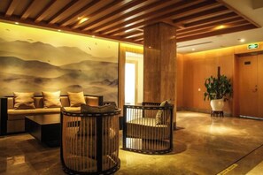 Un hall con techo de madera, un sofá, dos sillones de mimbre, una planta grande en maceta y una pared con un mural de paisaje montañoso.