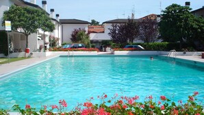 A swimming pool with a clear blue water, surrounded by a garden with red flowers, and buildings in the background.