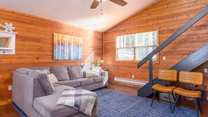 Ein Holzblock-Interieur mit einer Couch, Sesseln, einem Deckenventilator und einer Treppe.