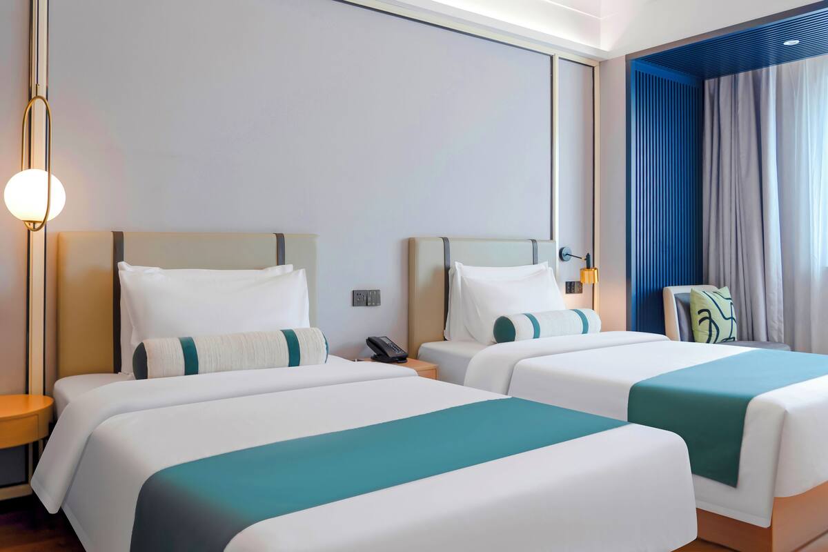 Een hotelkamer met twee bedden, een nachtkastje met een lamp, een telefoon en een raam met gordijnen.