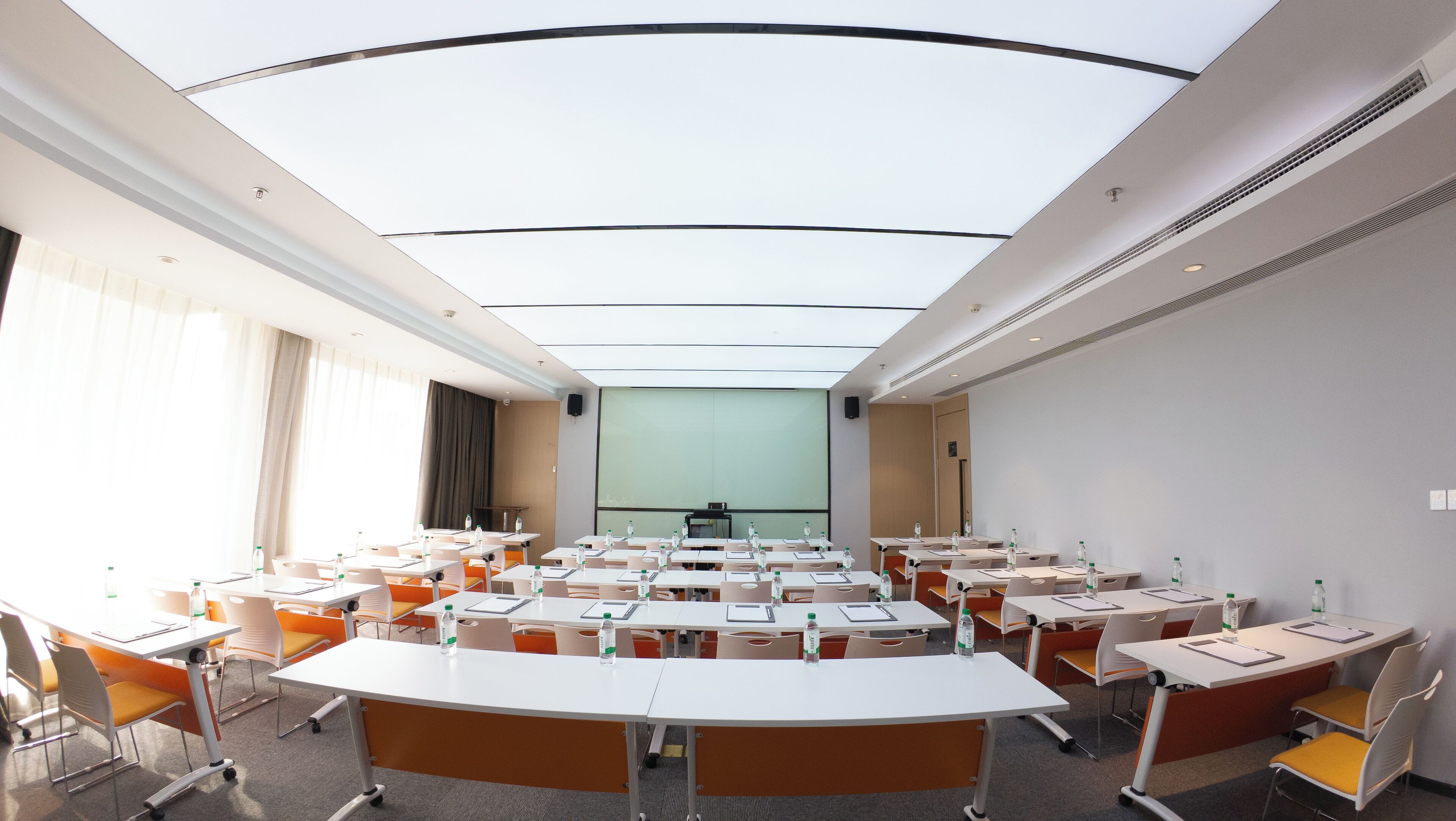 A conference room with a high ceiling, multiple tables with chairs, and a large screen at the front.