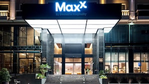Een hotelentree met een opvallend 'MaxX by Deutsche Hospitality'-bord, grote glazen deuren en een modern architectonisch ontwerp.