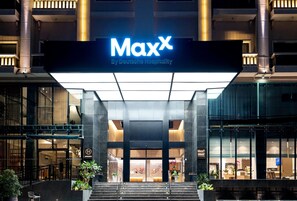 A hotel entrance with a prominent 'MaxX by Deutsche Hospitality' sign, large glass doors, and a modern architectural design.