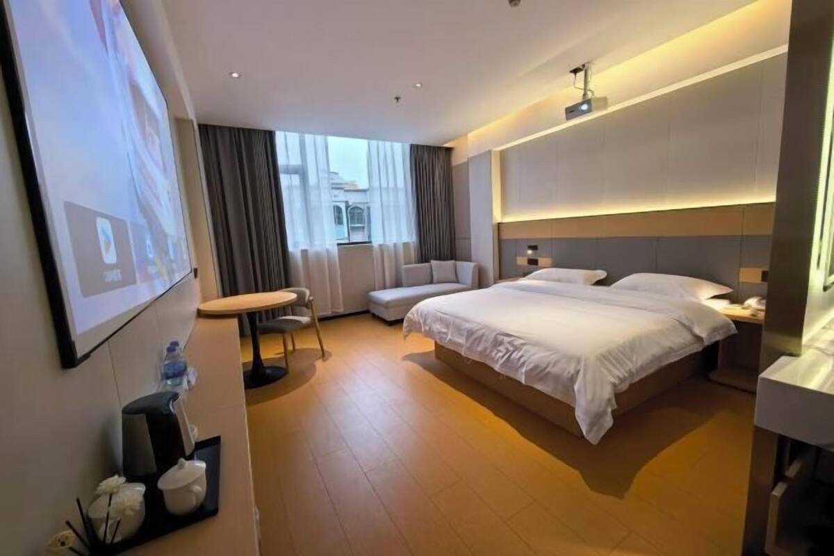 Habitación de hotel con una cama grande, un escritorio, una silla, una mesita, una pantalla de proyector y una ventana con cortinas.