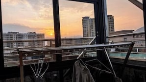 Une vue depuis une fenêtre, avec un étagère en métal où sont posés des verres et une veste, donnant sur un paysage urbain avec des bâtiments et un coucher de soleil.