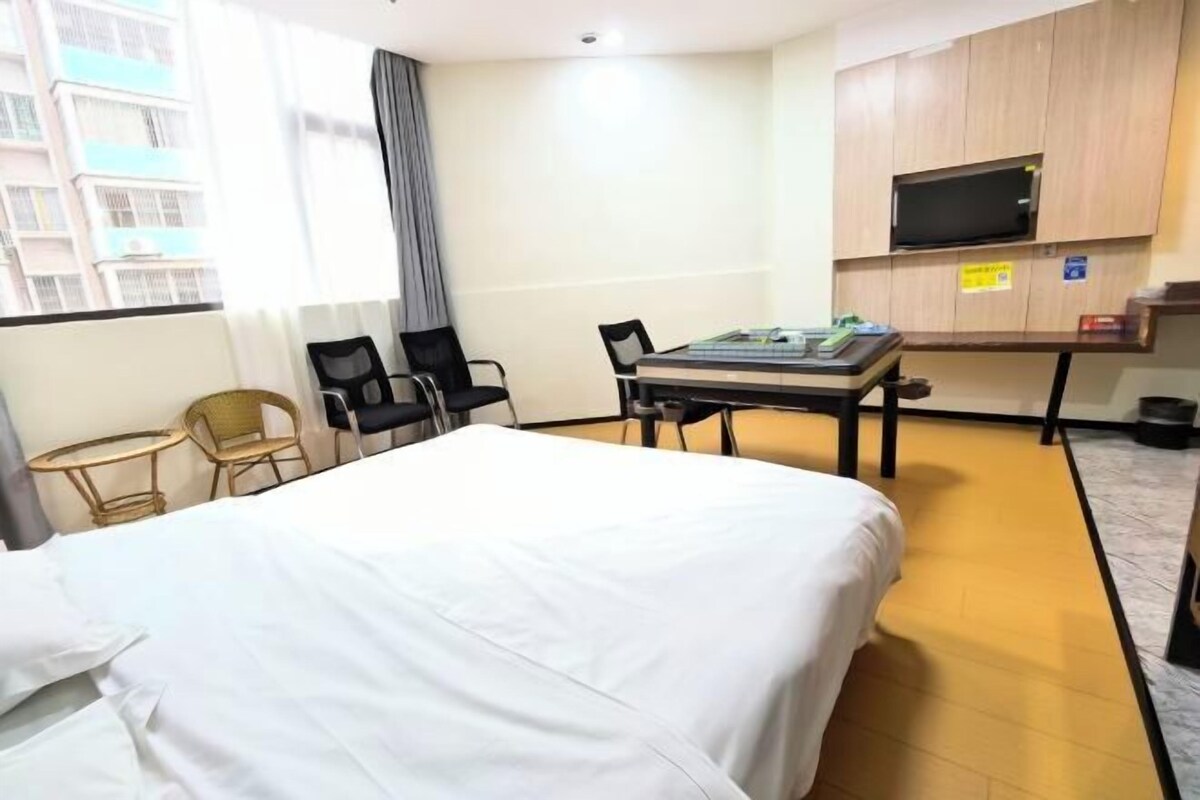A hotel room with a bed, a desk with chairs, a television mounted on the wall, and a window with a view of buildings.