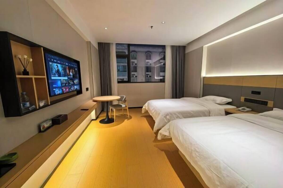 Habitación de hotel con dos camas, un televisor montado en la pared, una mesita y una ventana con vistas a edificios.