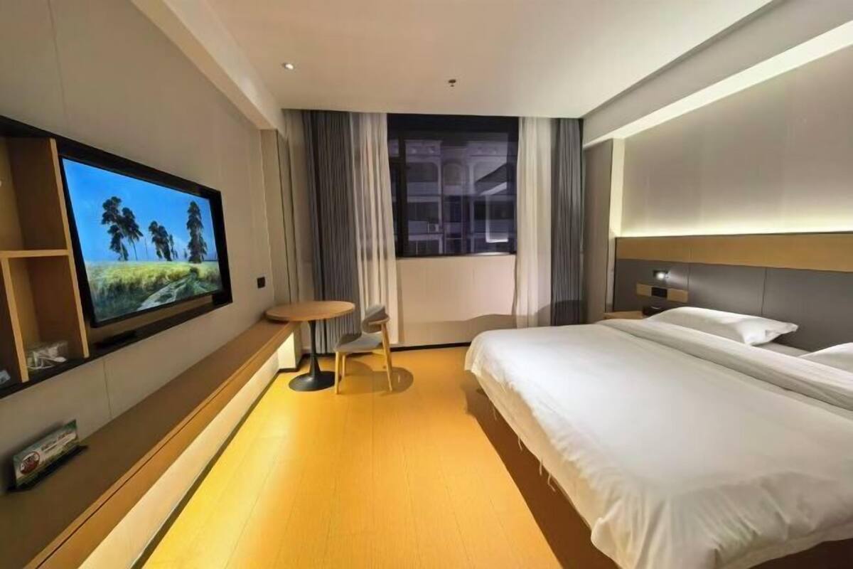Habitación de hotel con un televisor de pantalla plana grande montado en la pared, una cama con sábanas blancas, una mesita redonda con una silla y una ventana con cortinas.