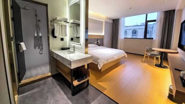 Une chambre d’hôtel moderne dotée d’un grand lit, d’une salle de bain séparée avec douche, d’une coiffeuse avec miroir et d’une petite table avec une chaise.