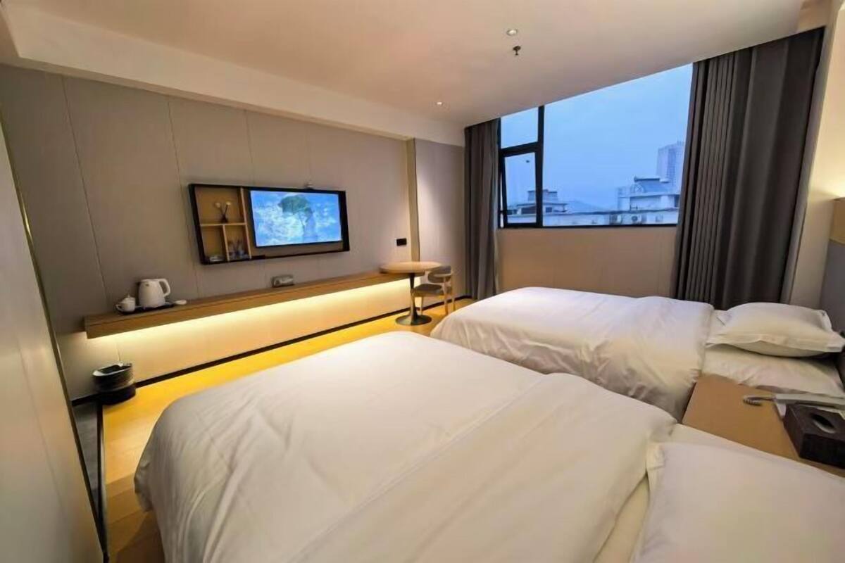 Habitación de hotel con dos camas, un televisor en la pared, un escritorio con silla y una ventana con vista a la ciudad.