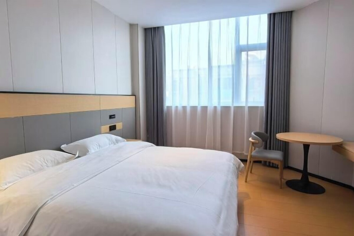 A hotel room with a large bed, a small table, a chair, and a window with curtains.
