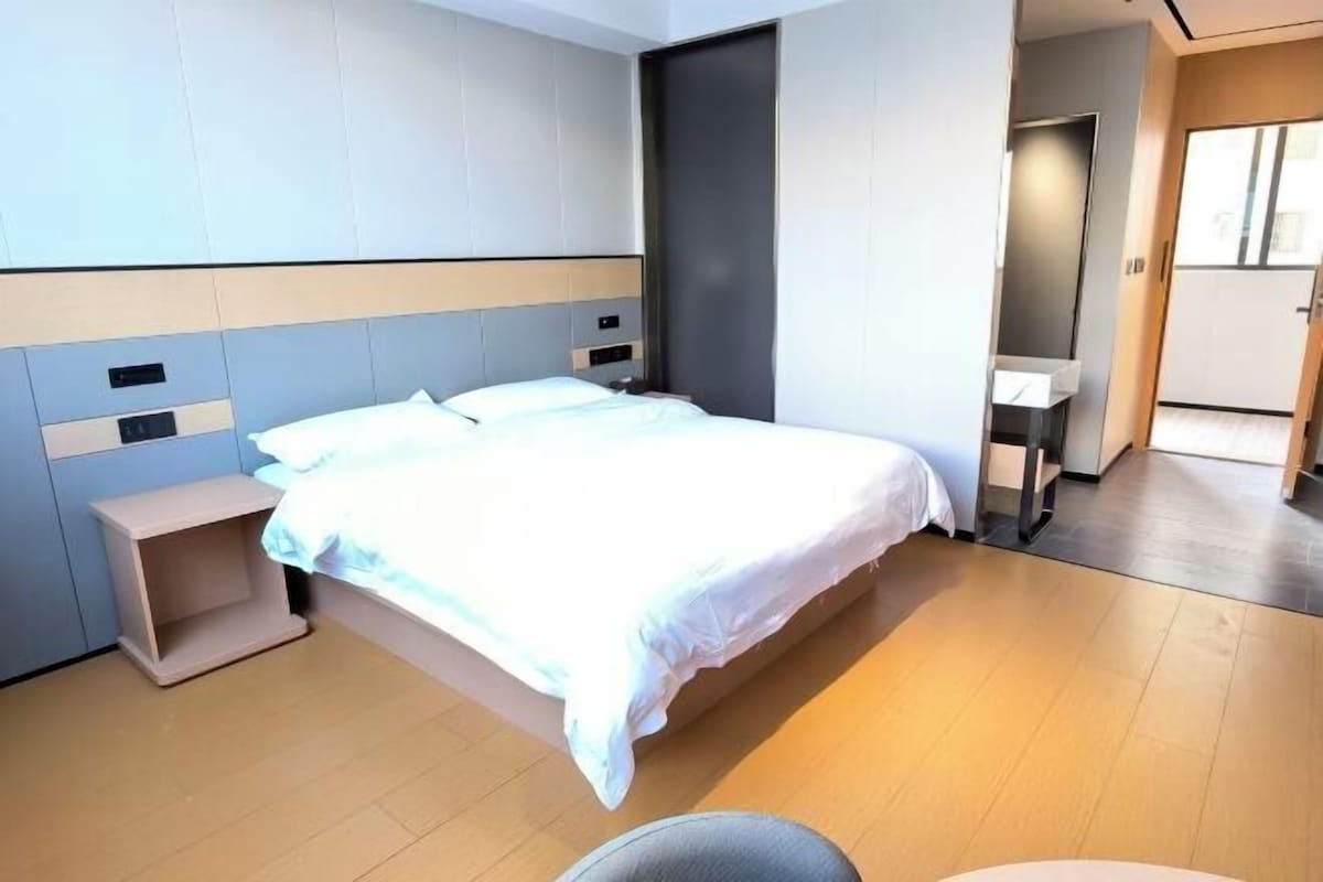 A modern hotel room with a large bed, a bedside table, a small desk, and a wardrobe.