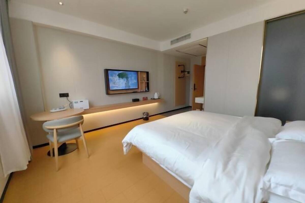 Una habitación de hotel con cama, escritorio con silla, televisión montada en la pared y un espejo.