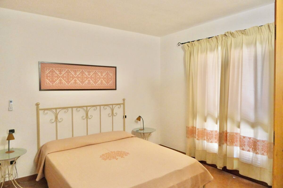 A bedroom with a bed, a small round table, a lamp, a framed picture on the wall, and a window with curtains.