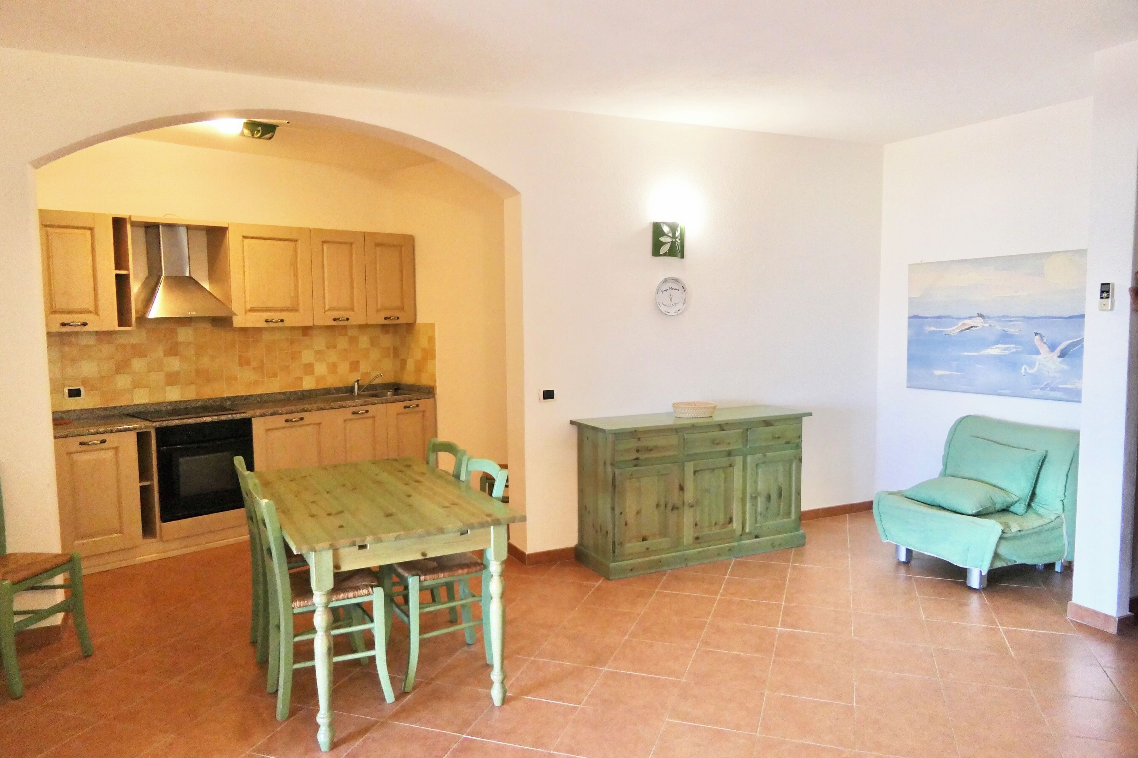 A kitchen with wooden cabinets, a dining table with chairs, a green sofa, and a painting on the wall.