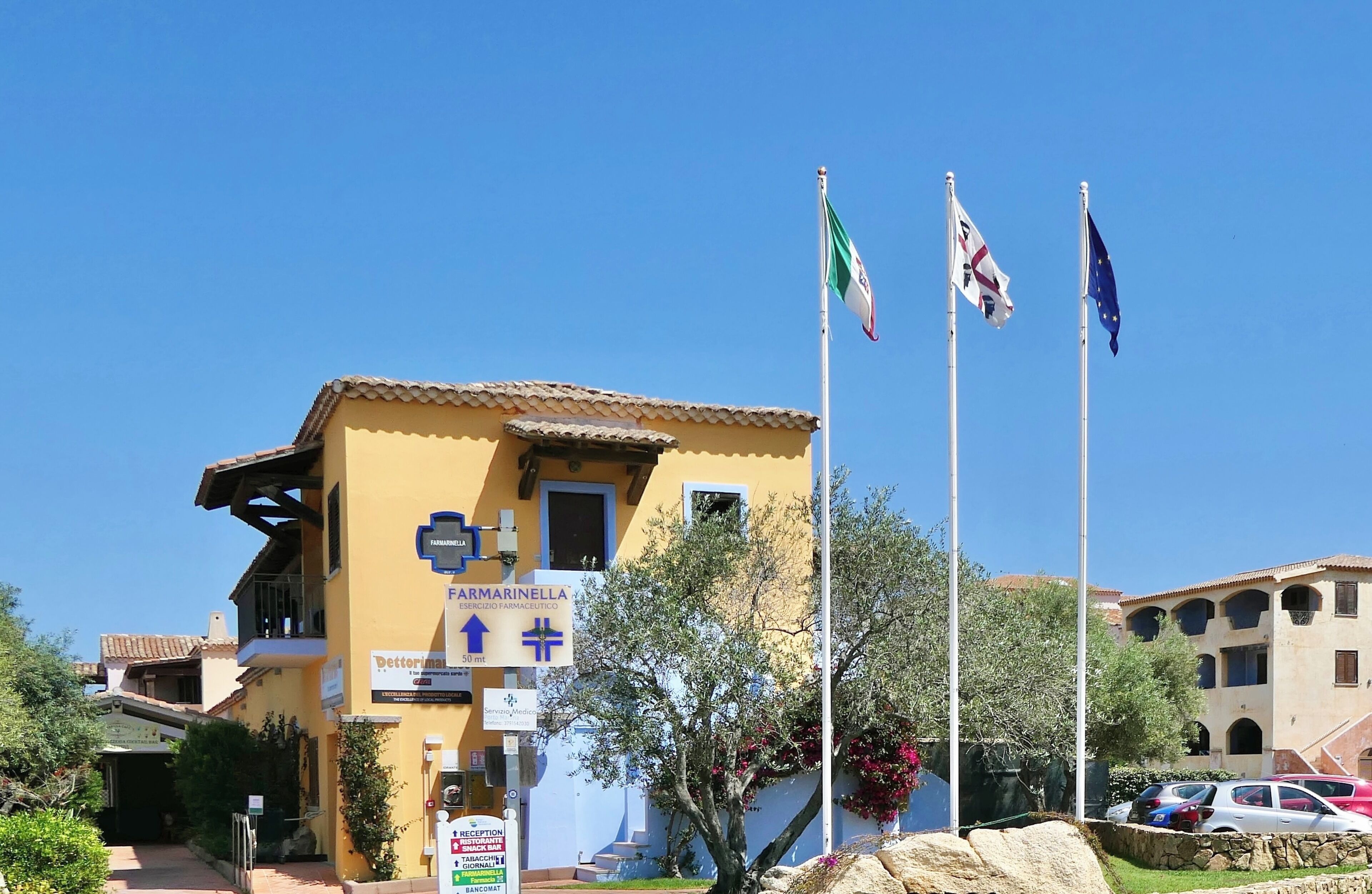 A yellow building with a sign for 'FARMARINELLA ESSICERIO FARMACEUTICO' and flags, including the Italian flag.