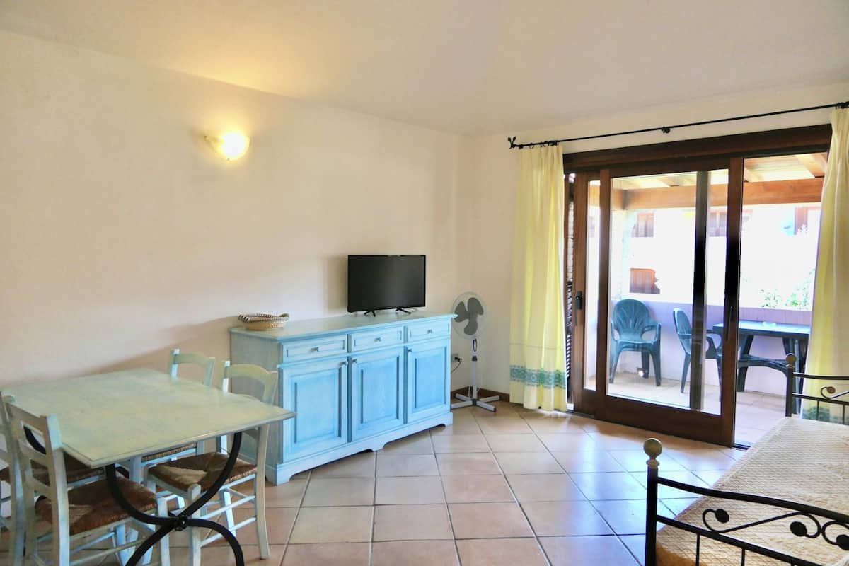 A room with a dining table, chairs, a TV, a fan, and a sliding glass door leading to an outdoor area with chairs.