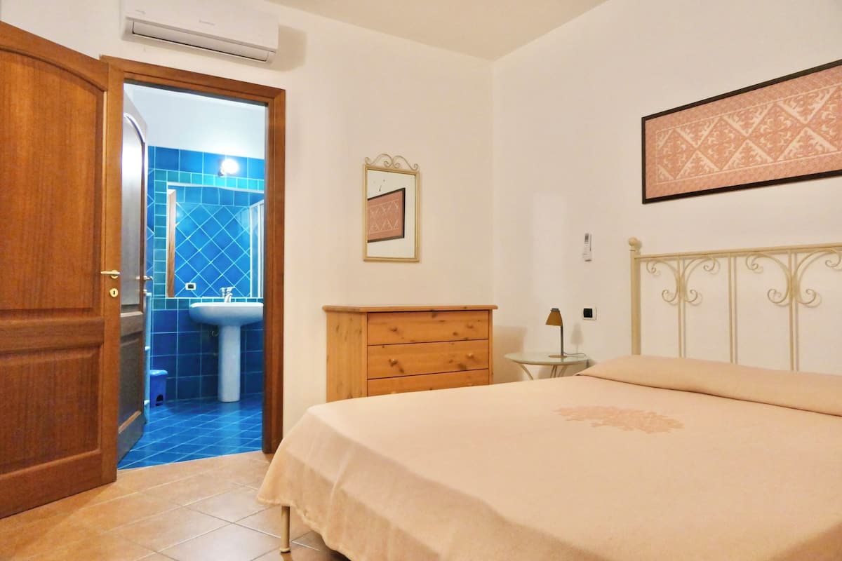 A bedroom with a bed, a wooden dresser, a nightstand, a framed picture on the wall, and a bathroom visible through an open door.
