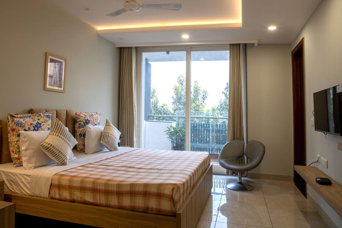 A bedroom with a bed, a chair, a TV, and a balcony with a view of trees.
