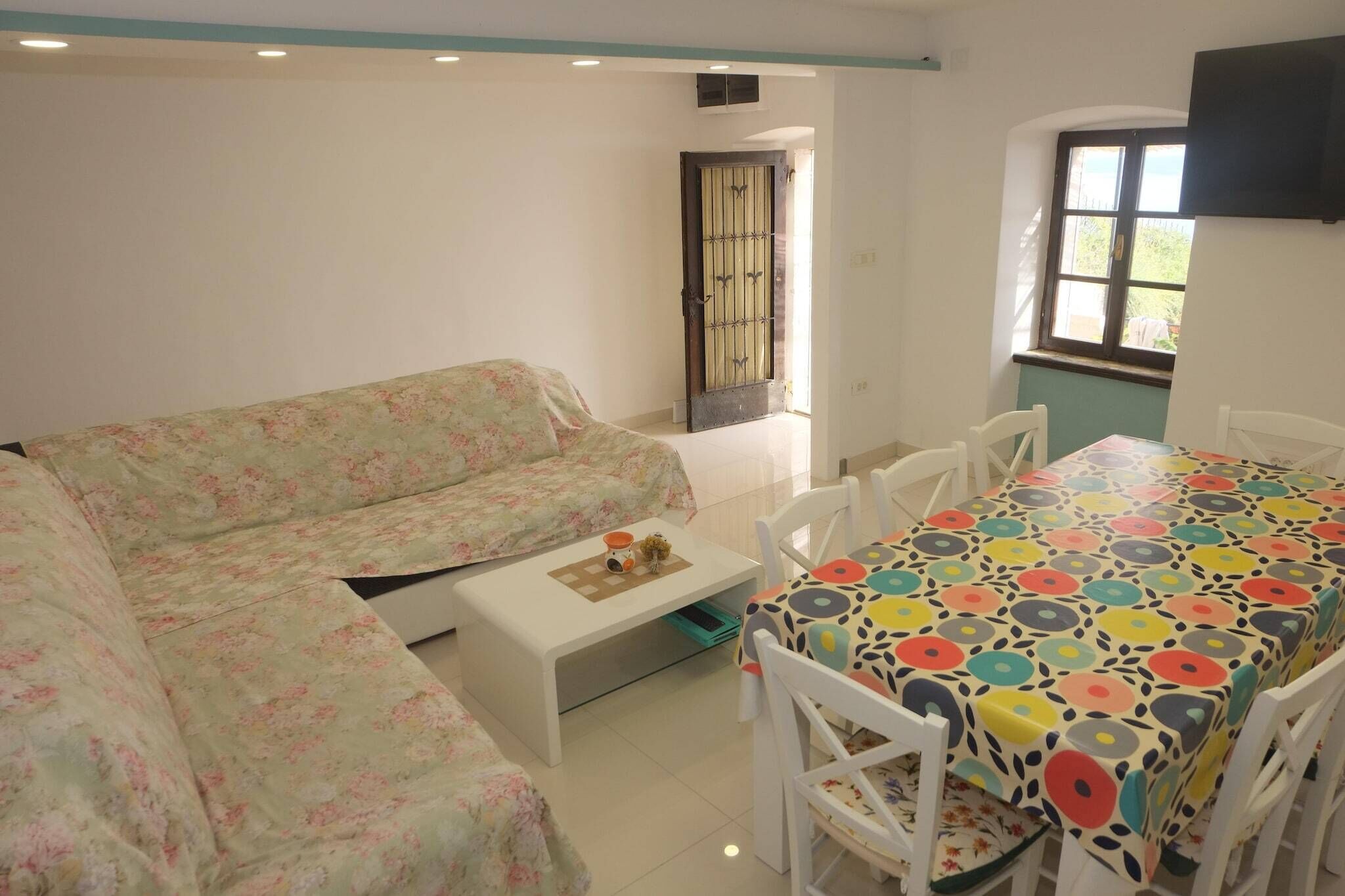 Une pièce avec un canapé à motifs floraux, une petite table blanche sur laquelle se trouve un plateau, et une table à manger recouverte d’une nappe colorée, avec des chaises blanches.