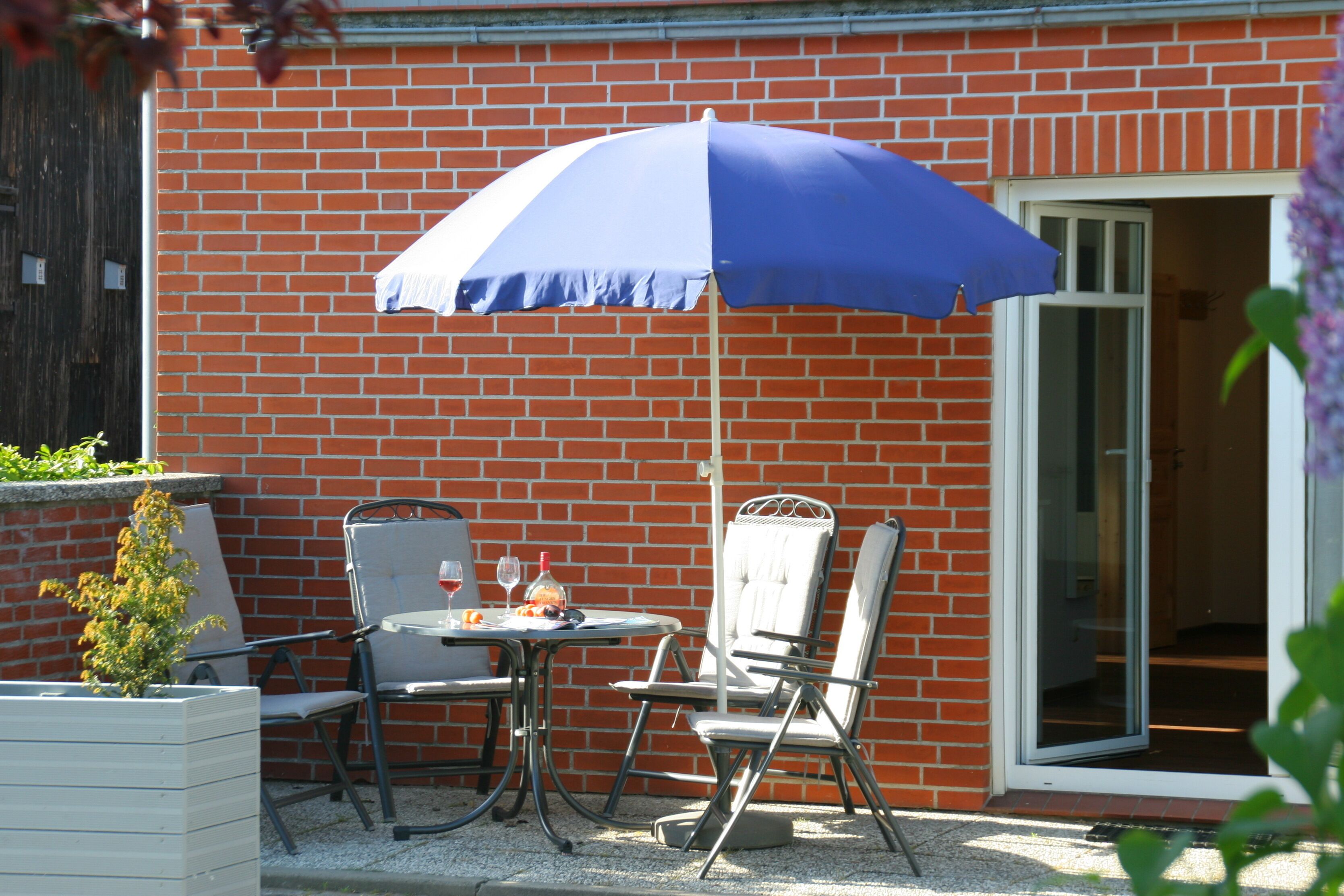 Een terras met een tafel, stoelen en een blauwe parasol, tegen een bakstenen muur.