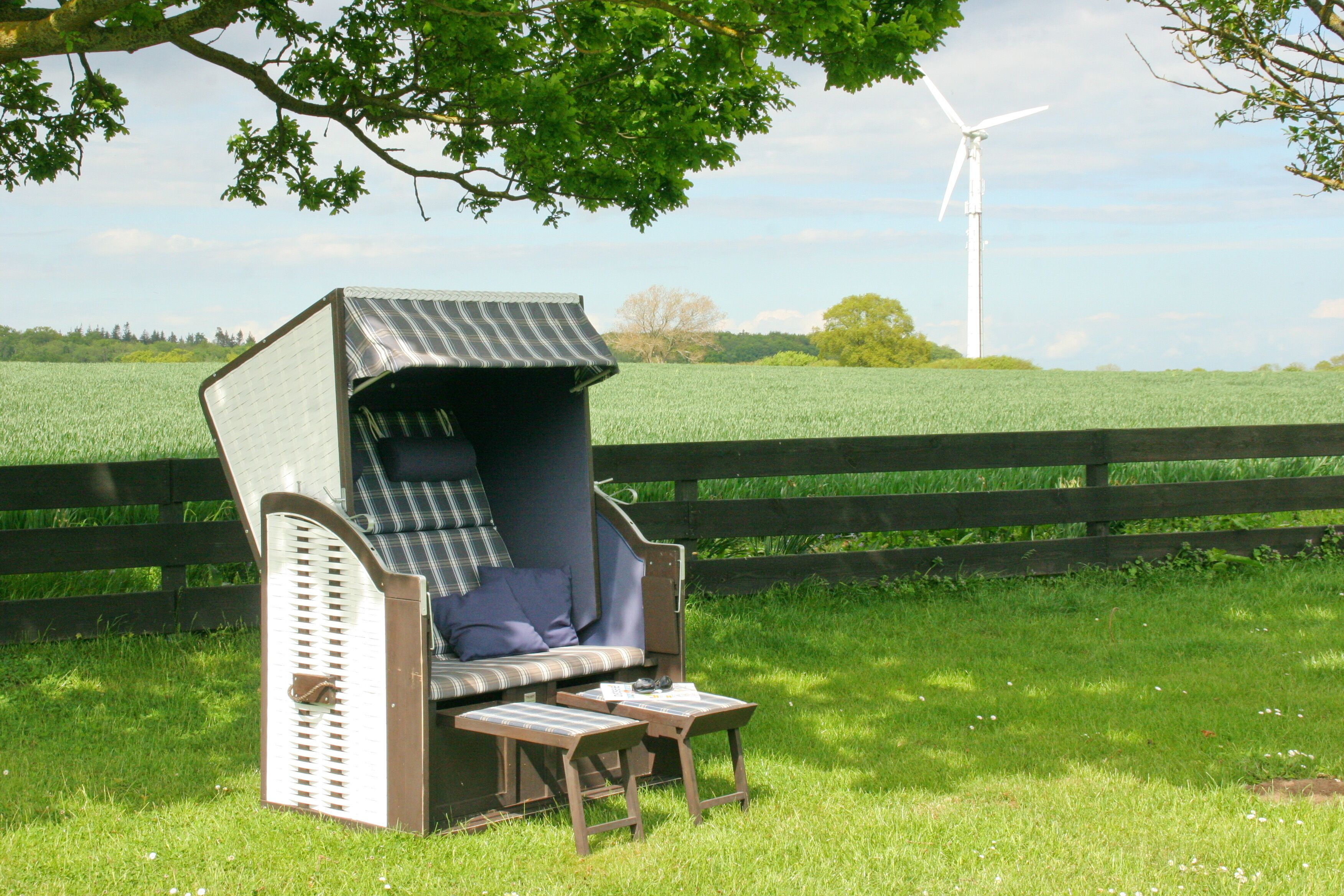 A cozy outdoor seating area with a high-backed chair and a small table, set against a backdrop of a green field and a wind turbine.
