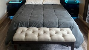 Een slaapkamer met een modern design, met een houten hoofdbord, een grijze spreideken en een bankje.