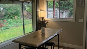 Een eetruimte met een tafel en twee krukken, een vaas met bloemen, een raam met uitzicht op groen, en een plafondlamp.