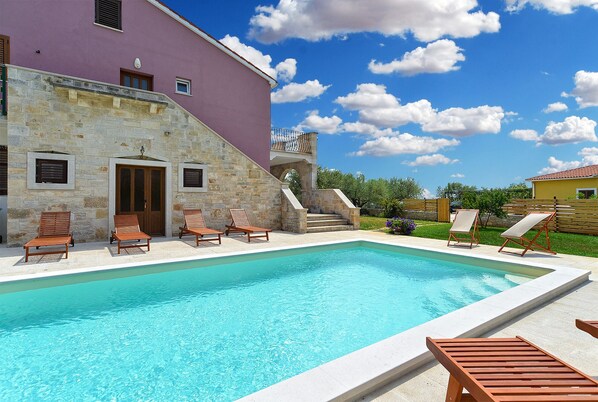 A swimming pool with wooden loungers, a stone building with a purple facade, and a clear blue sky with clouds.