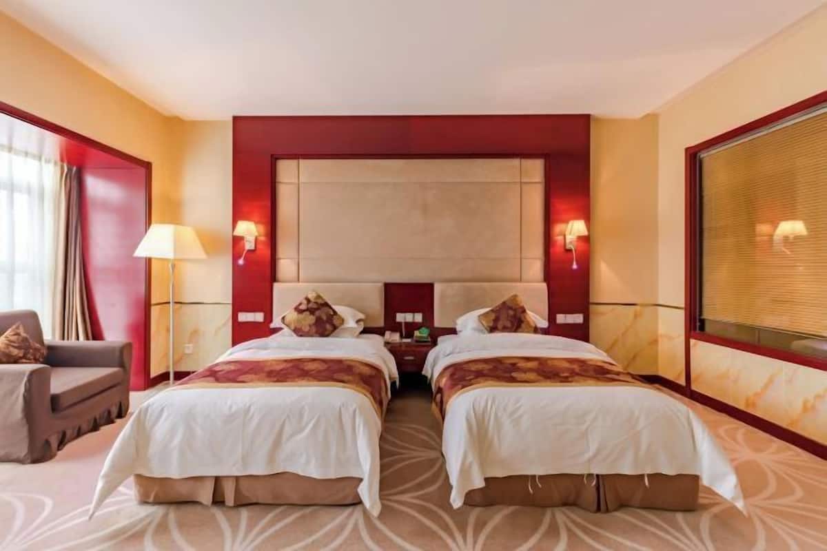 A hotel room with two beds, a sofa, a lamp, and a window with blinds.